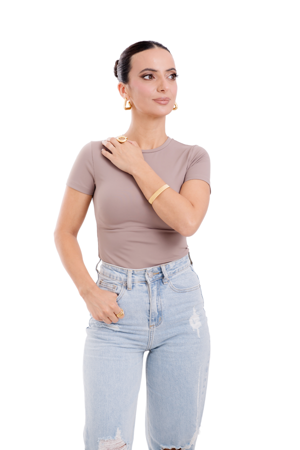 Fashionable woman wearing a fitted taupe top, ripped denim jeans, and bold gold jewelry, showcasing a chic and minimalist style