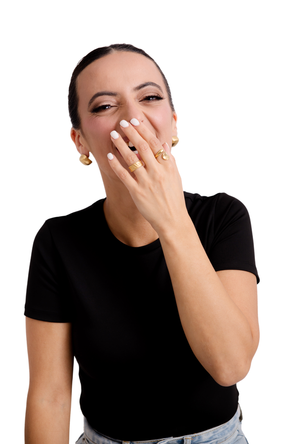Woman in a black top laughing joyfully, wearing bold gold rings and earrings, exuding confidence and playful elegance.