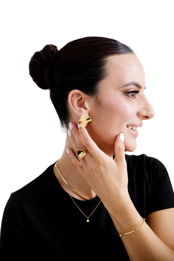 Elegant woman wearing contemporary gold jewelry, including bold sculptural earrings, a matching gold ring, a delicate necklace, and a bracelet, styled with a black top and a sleek updo hairstyle.
