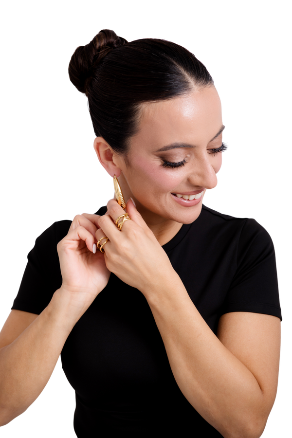Smiling woman in a black top adjusting gold statement earrings and showcasing bold gold rings, highlighting modern and elegant jewelry.