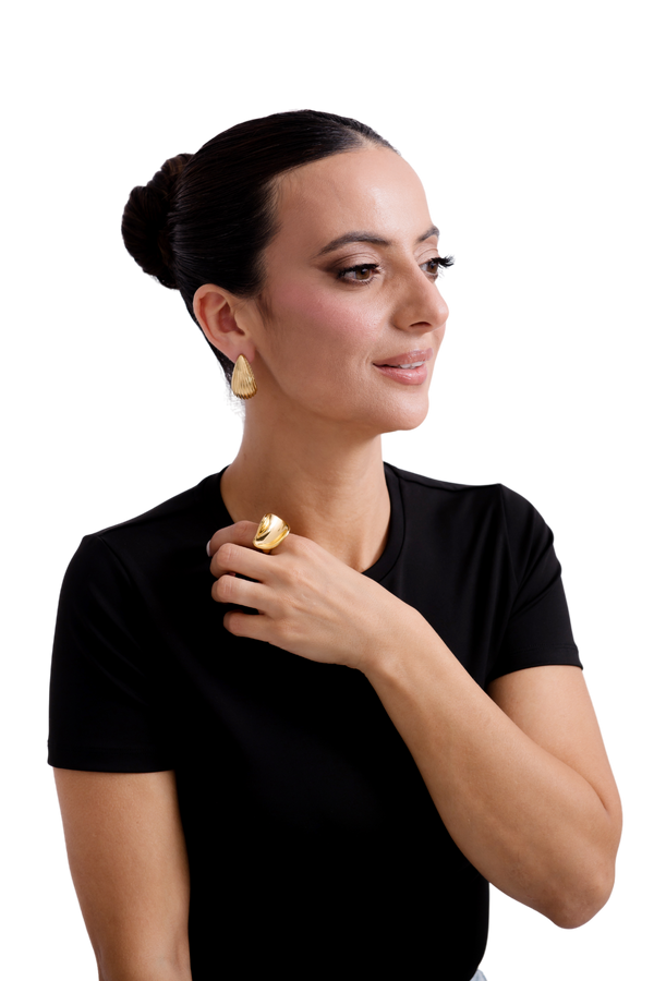 Stylish woman wearing gold statement jewelry, featuring leaf-shaped earrings and a bold gold ring, paired with a sleek black top and an elegant updo hairstyle.